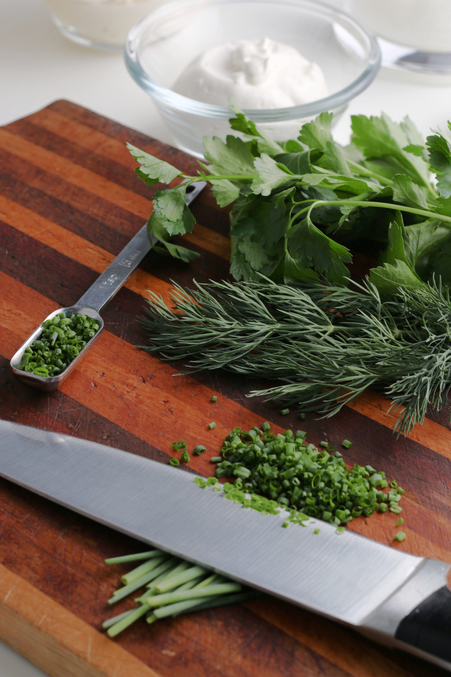 Fresh herbs used to make ranch dressing sit on a wooden cutting board with a chopping knife.