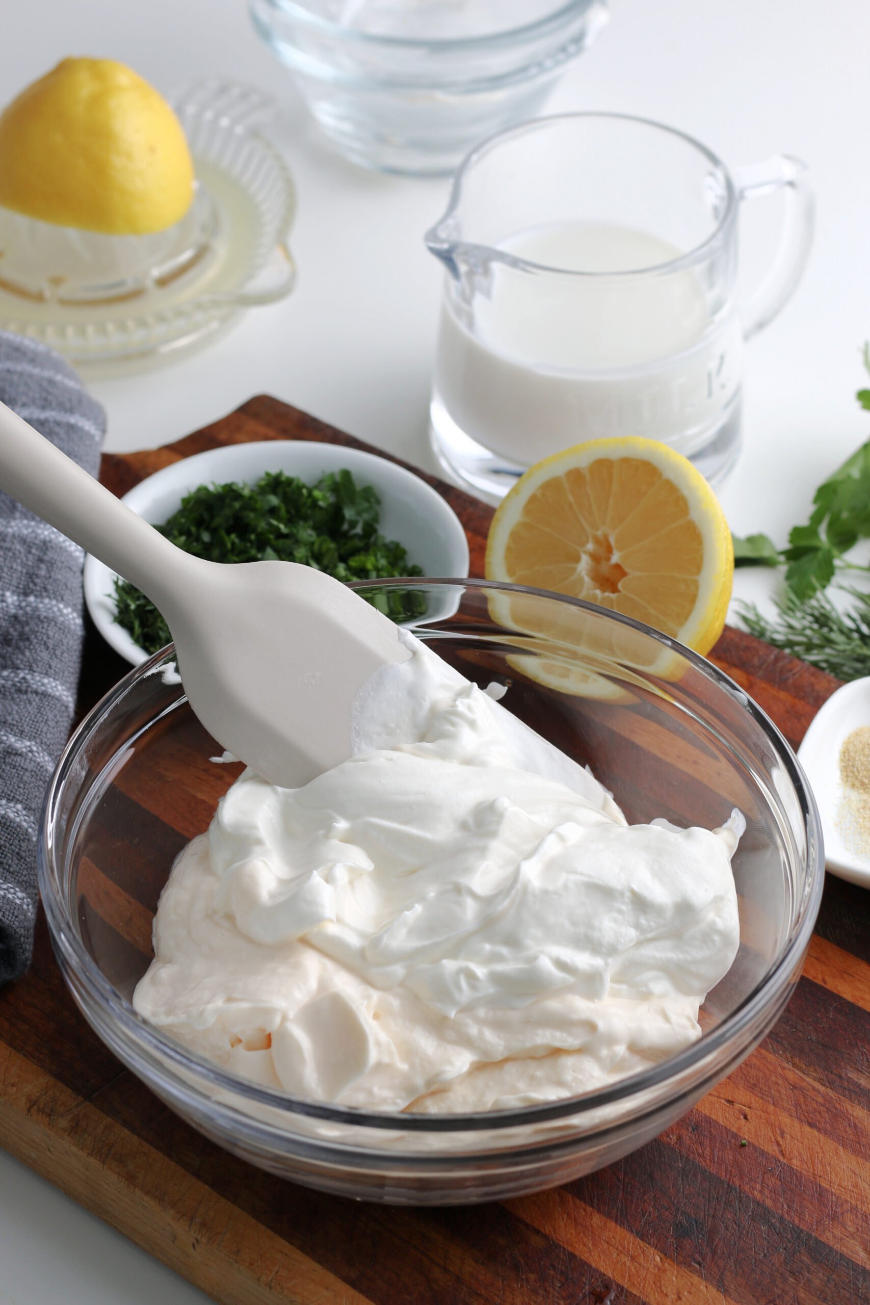 A close-up of the base ingredients for ranch dressing with a white spoon inside the glass bowl. The remaining ingredients are in the background.