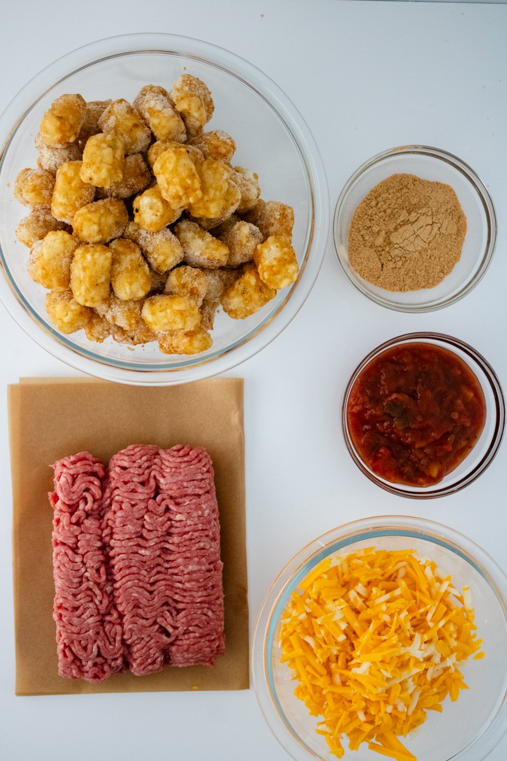 The ingredients for taco tot casserole sit in glass bowls on a white countertop. The raw meat sits on top of brown parchment paper.