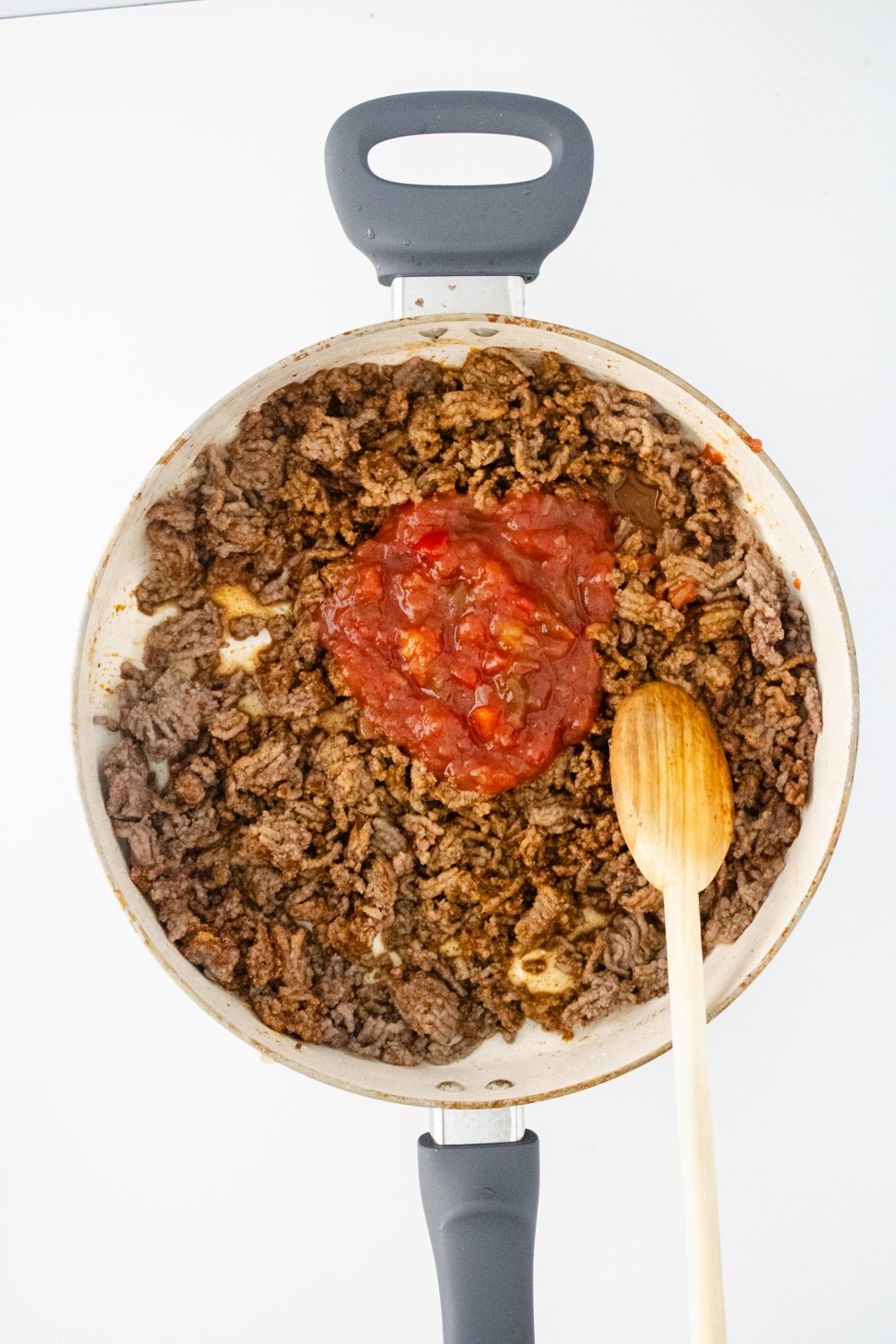 A pan is filled with cooked ground beef that is topped with salsa. A wooden spoon is resting on the side of the pan.